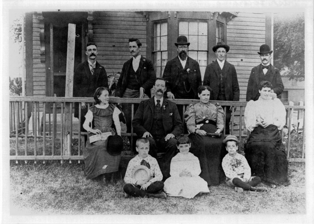 Men, women in children in fancy dress posed on the porch of a boarding house.