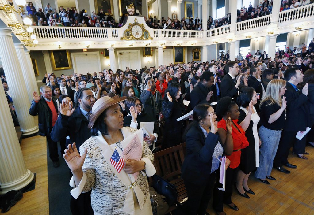 Many men and women stand with their right hand over their heart, holding an American flag and naturalization papers, in the process of taking their oath of citizenship. People are gathered on the floor of Faneuil Hall, but some too in a second-floor gallery.