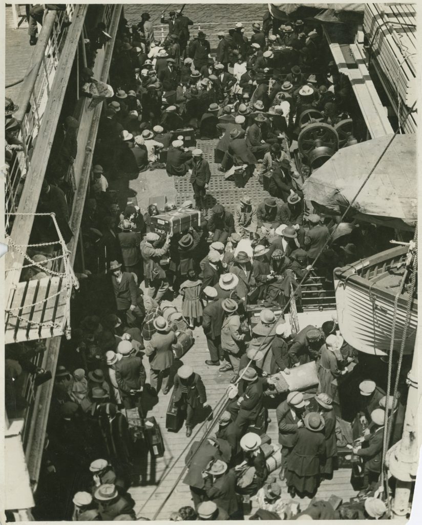 A bird's eye view of a deck of a ship bringing immigrants to Boston. Many immigrants are crowded on the deck, either standing or sitting in groups.