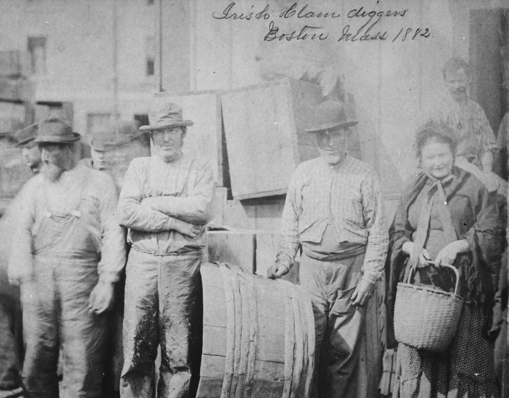 A photograph of six men and one women wearing work clothes. In the center of the photograph is a large barrel and the woman holds a basket. On the top of the photograph, it says "Irish Clam Diggers, Boston, Mass, 1882" in script.