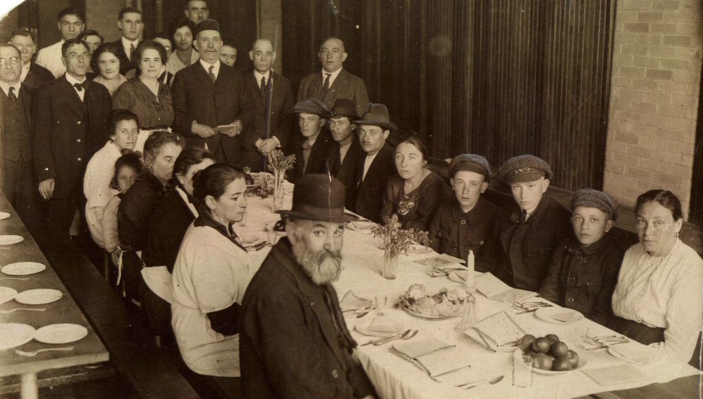Fourteen men, women, and children are seated at a table in preparation for Passover seder. Many men and women stand t the head of the table. The table is decorated nicely with flowers and a white tablecloth.