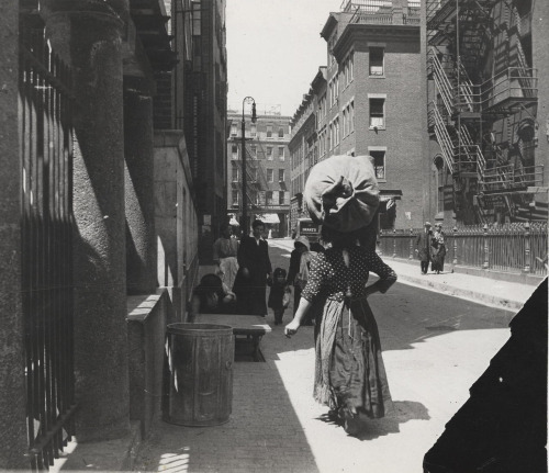 An Italian woman walks down a busy street in the North End with a large filled bag carried on her head.