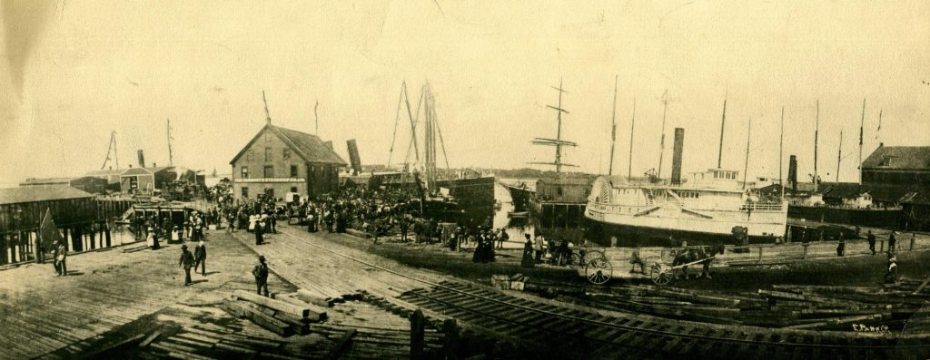 A train track begins near a dock and various ships. Many people are gathered near the dockhouse.