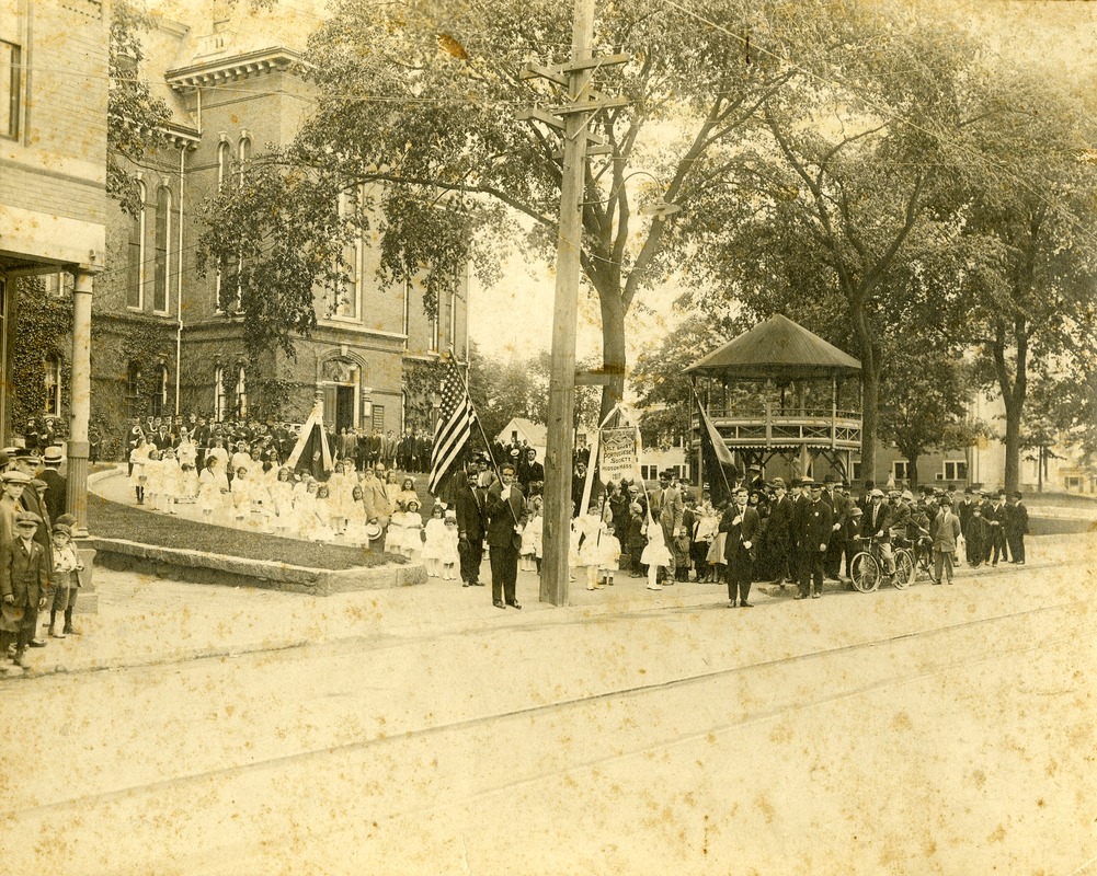 A group of men, women, and children are gathered in front of a building to take place in the Trinity Sunday procession. At the front is a man holding an American flag. Behind him are many young children all dressed in white. Behind the children are the adults, dressed nicely.
