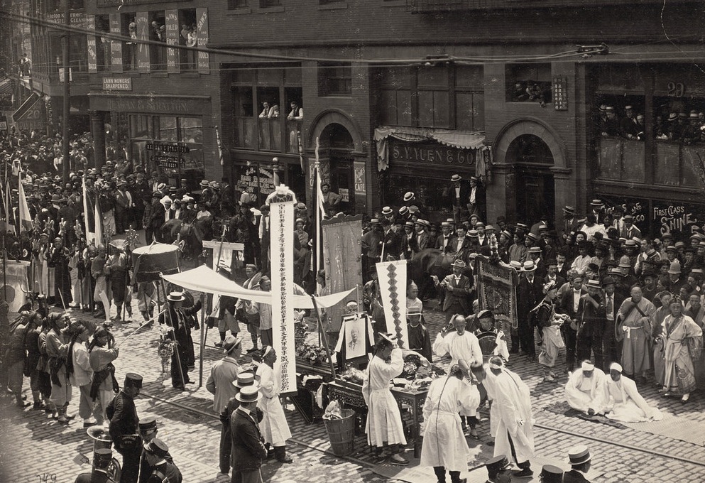 A traditional Chinese funeral takes place in the middle of Harrison Street. On either side of the street are hundreds of men watching and taking place in the funeral.