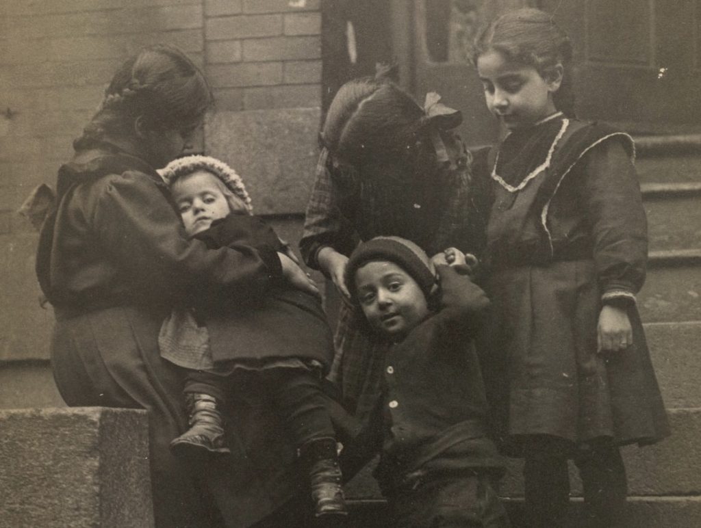 Five young children are gathered together on the steps of a building. Three young girls care for two younger toddler-aged children.