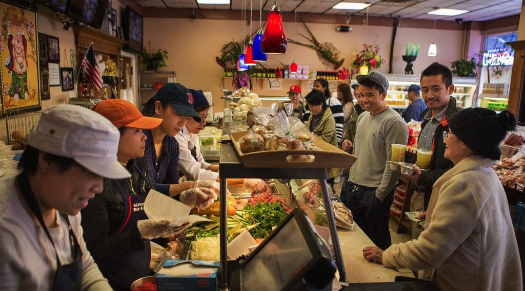 A line forms at the counter of Banh Mi Be Le. Ten people are waiting in line or are in the process of ordering at the counter as four women work behind the counter to fulfill their orders.