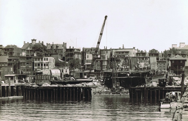 The construction of a ferry in the water using a crane and other tools. Behind the construction are various multi-story buildings.