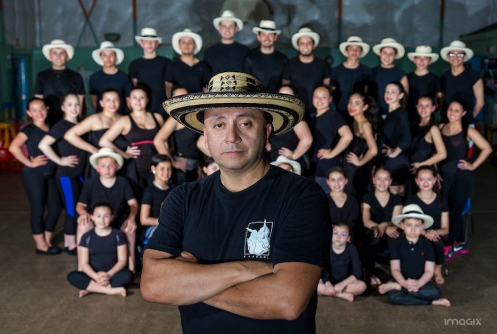 A man in a traditional sombrero vueltiao stands near the camera with his arms crossed. Behind him are four rows of young children, the boys all in a similar hat.