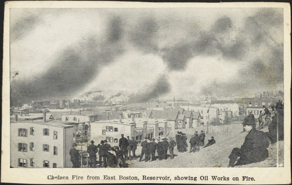 Postcard captioned "Chelsea Fire from East Boston, Reservoir, showing Oil Works on Fire." Many men dressed in hats and suits stand looking out towards the fire and the street of residential apartment buildings below. In the distance is the fire along with large plumes of smoke.