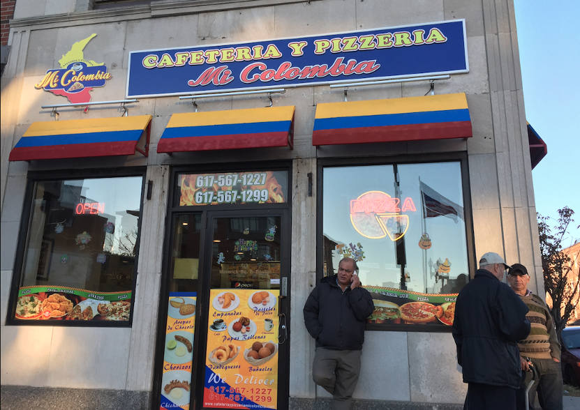 A photograph of the facade of "Cafeteria y Pizzeria Mi Colombia." The signage and awnings evoke the Columbian flag. The door and windows illustrates some of their menu items. Three men stand outside of the store.