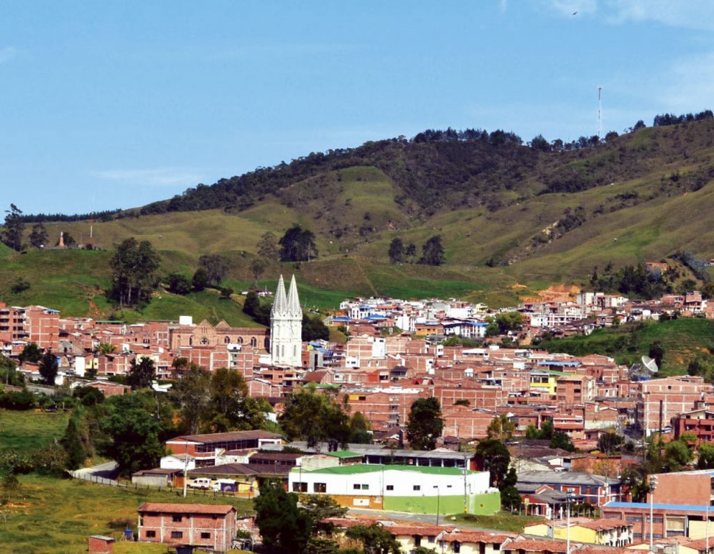 A photograph of a bustling town located in the foothills of a mountain. There are many brick buildings located close together growing up the hill, along with a large white spire in the center of the town.