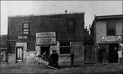 A photograph of two businesses next to each other. On the left is a scarp dealer with a wagon and two men standing out front. On the right is a poultry shop.