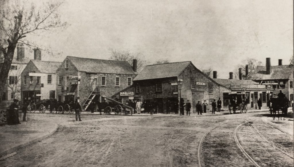 A photograph of an intersection between two dirt roads. Horse-drawn carriages line one of the streets, in front of commercial businesses. The buildings are are all generally two or three stories and are in a colonial style.