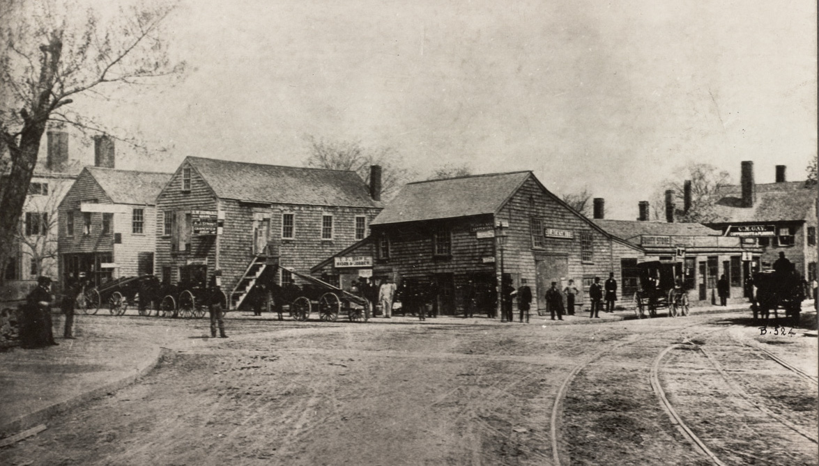 A Photograph Of An Intersection Between Two Dirt Roads. Horse-drawn Carriages Line One Of The Streets, In Front Of Commercial Businesses. The Buildings Are Are All Generally Two Or Three Stories And Are In A Colonial Style.