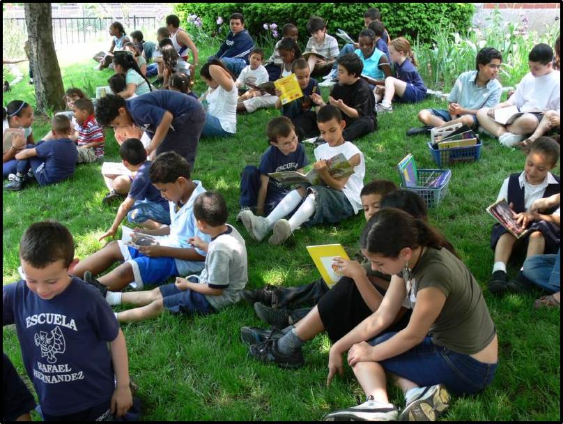 Pairs of children sit on the ground reading various children's picture books from a basket. Many of the young children wear a shirt that reads "Escuela Rafael Hernandez."