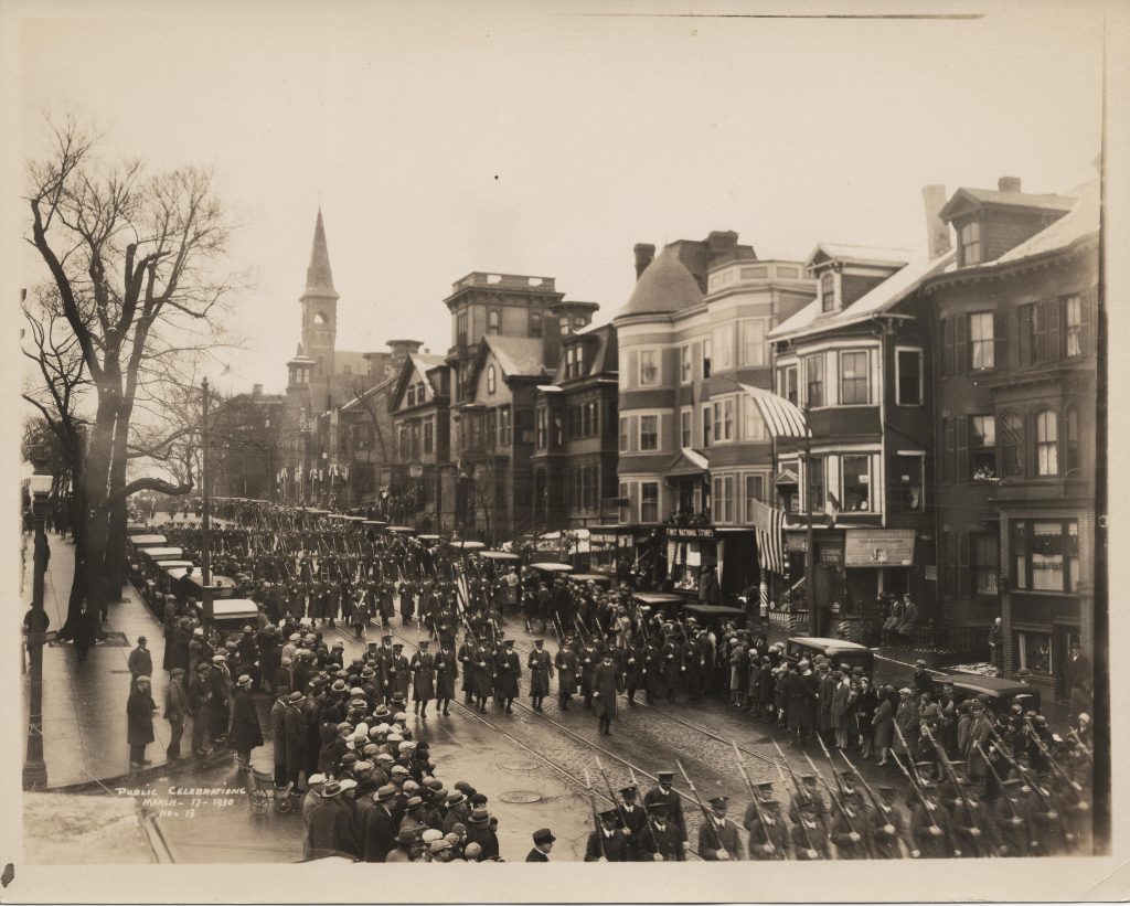 A photograph of a parade with crowds lining either side of the street. Uniformed men holding rifles walk in the parade, separated into groups. There are multi-story mixed-use buildings on the side of the street.