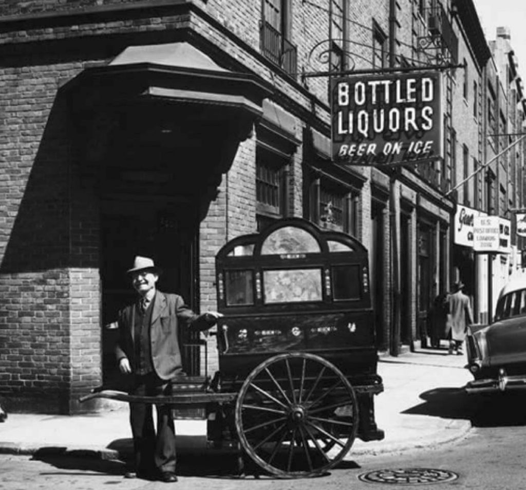 A man smiles standing next to his portable organ with his hand resting on the side of the machine. The instrument is on wheels and has two handles on either side to transport. Behind the man is a brick liquor store.