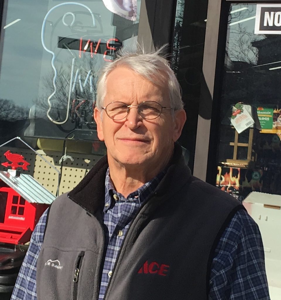 An older man wearing glasses, a plaid button-down and an Ace hardware vest looks into the camera. He stands in front of a hardware store with the window displays in the background.