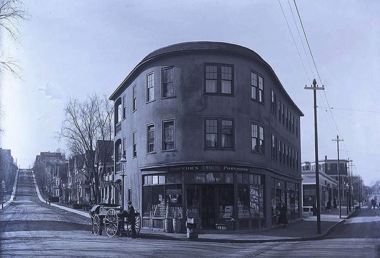 A photograph of Belgrade Avenue and Birch Street. On the corner, in the center of the photograph, is a curved building that is three-stories. On the first floor is a grocer with a sign that reads "T.M. Nulty: Groceries, Provisions." In front of the store is a man with a horse-drawn wagon.