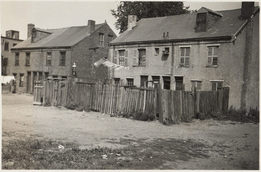 A photograph of two two-story houses in a colonial style. The house in the center of the photograph has backyard fenced off by an old wooden fence with a clothesline in the air. Behind the houses is a dirt road.