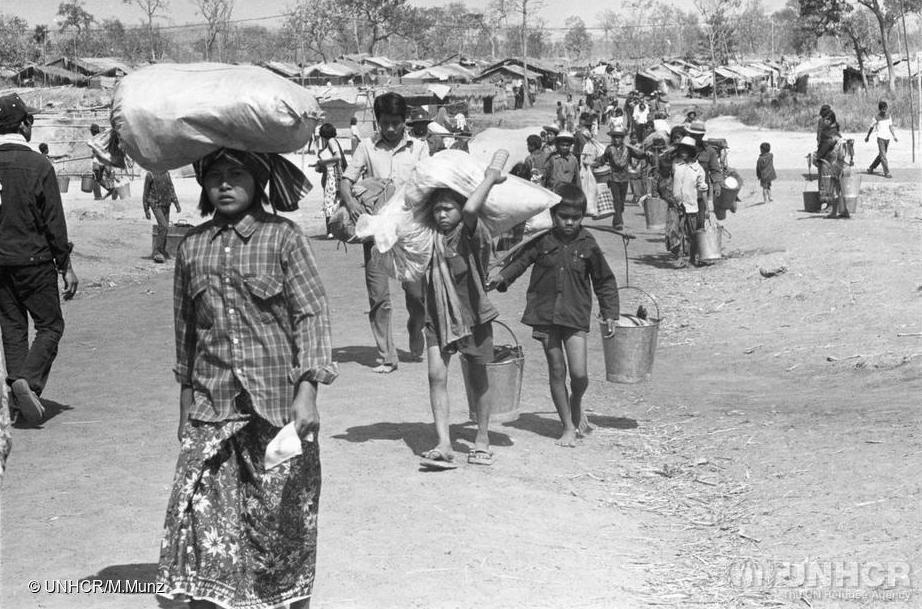 Three Cambodian children walk, carrying various belongings on their heads and in buckets on either side. Behind them is a long line of other refugees following suit. In the background is an area with many small buildings.