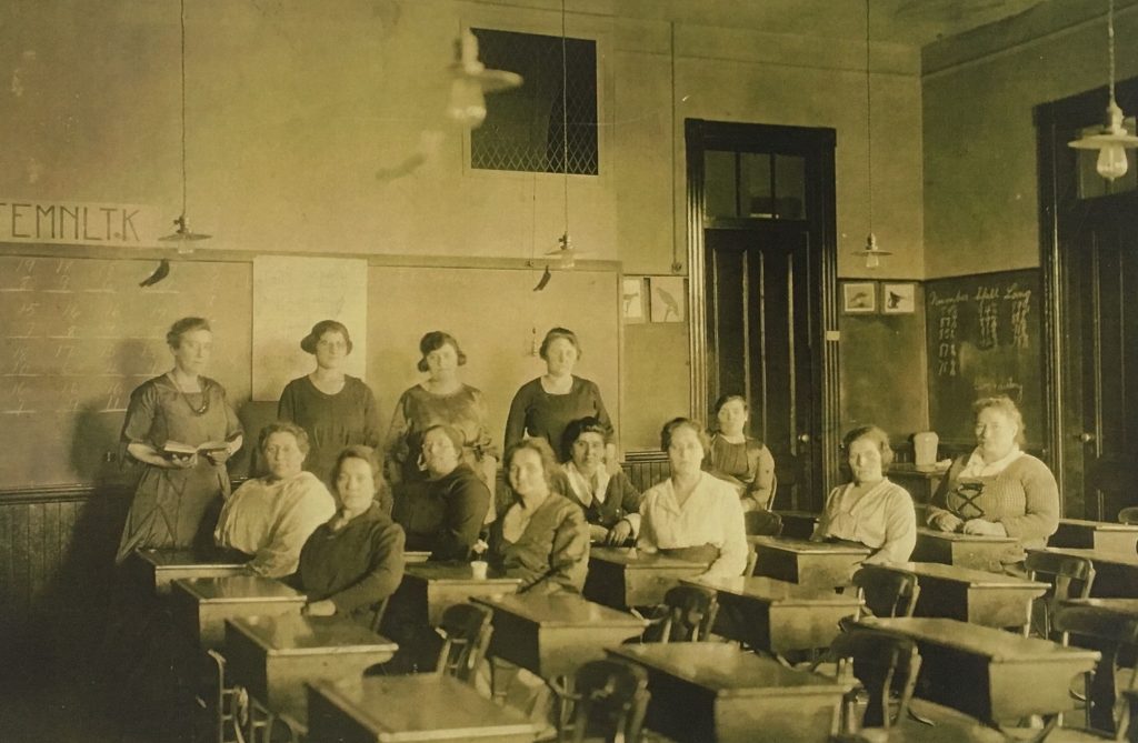 A classroom with six rows of four desks. Four women stand behind the desks, one holding a book. Nine women are seated in the desks towards the back of the classroom. Behind the women are chalkboards with mathematic on them.