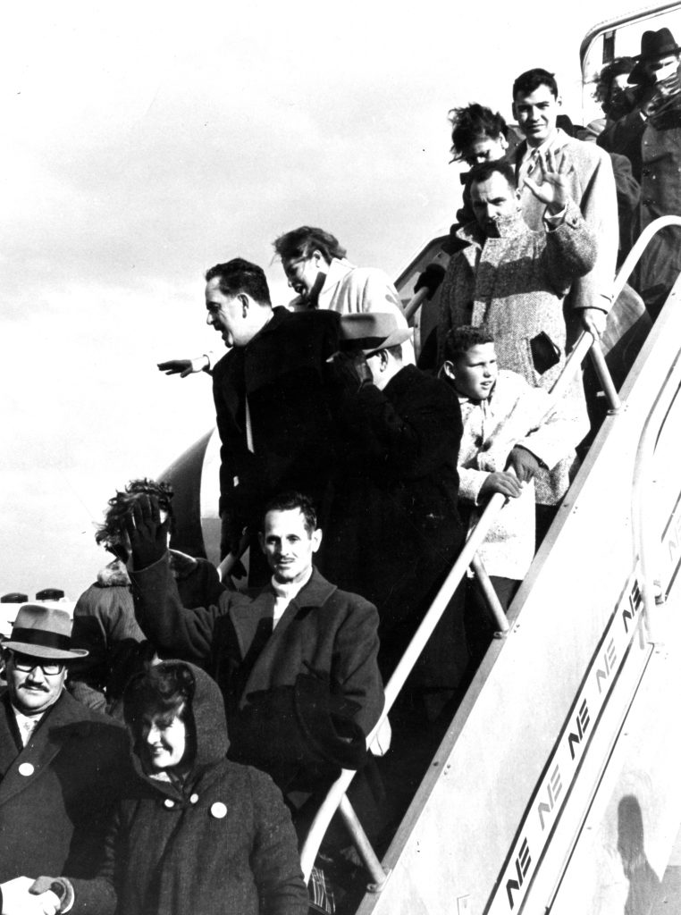 Fifteen men, women, and children getting of an airstair and arriving at Logan airport. Some of the refugees wave to the camera or wave elsewhere. All are dressed warmly, in large coats.