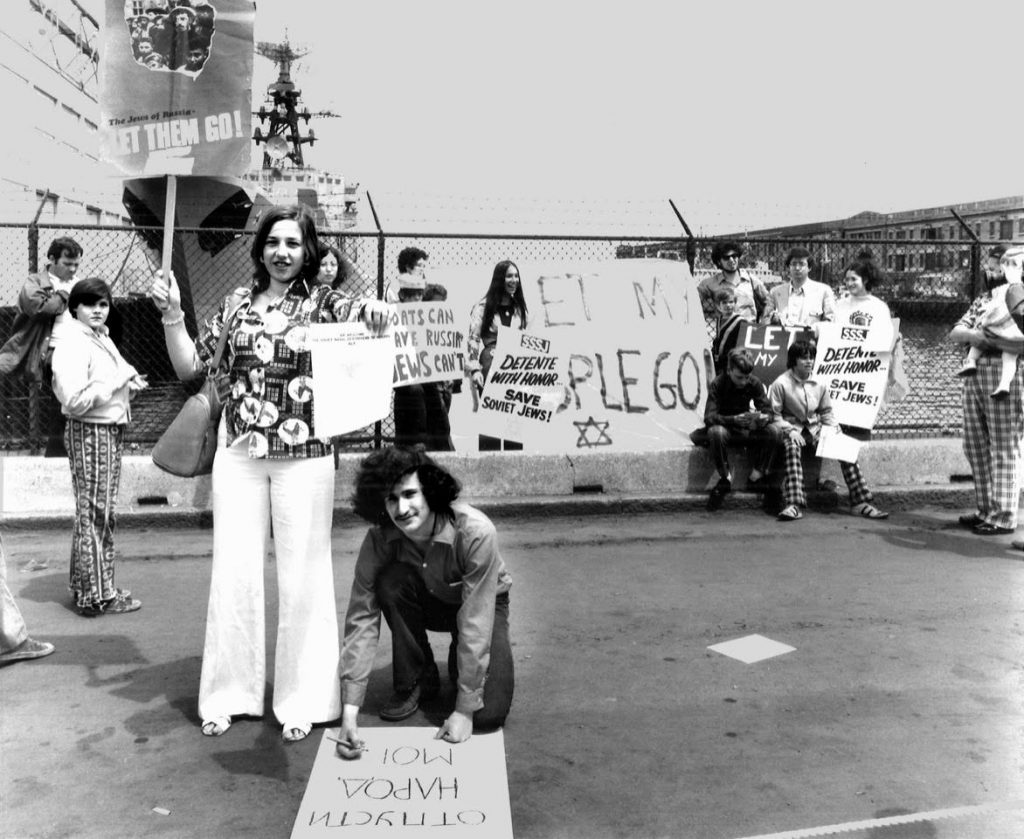 Seventeen people hold posters and protest the arrival of a large Soviet ship, which is in the background of the photograph. Many of the posters read "Detente with Honor, Save the Jews." In the front of the photograph is a man writing on his own poster in Russian, and a woman standing with a sign that reads "The Jews of Russia-- let them go!"