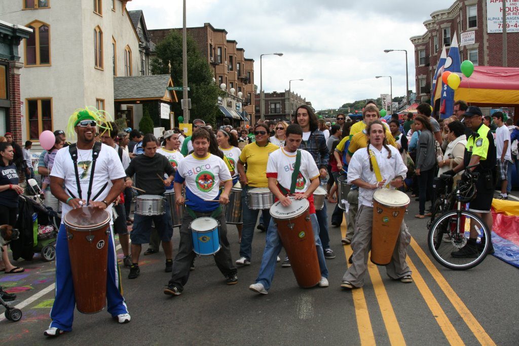 A bustling street parade. In the middle of the street, four men play drums with their hands. Behind the men are various people playing snare drums. There are many spectators on either side of the street.
