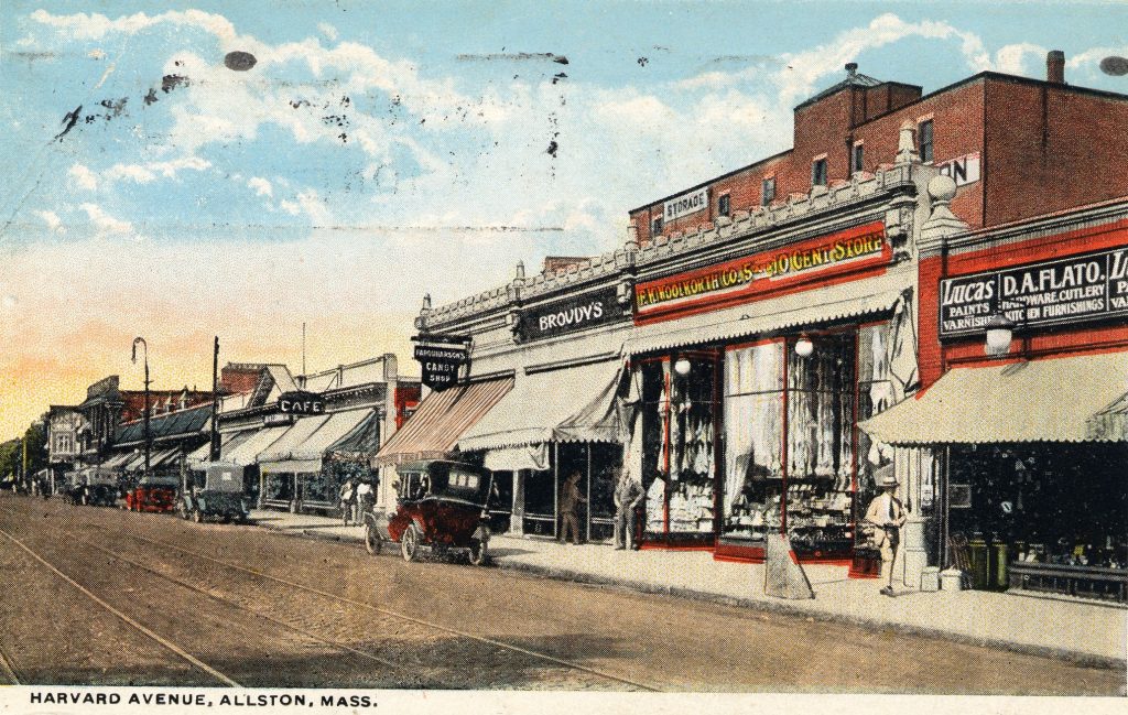 A postcard of "Harvard Avenue, Allston, Mass." There is a street on the left of the postcard with lines for a streetcar. On the side of the street are numerous businesses, such as a ten cent store, candy store, and cafe, along with automobiles.