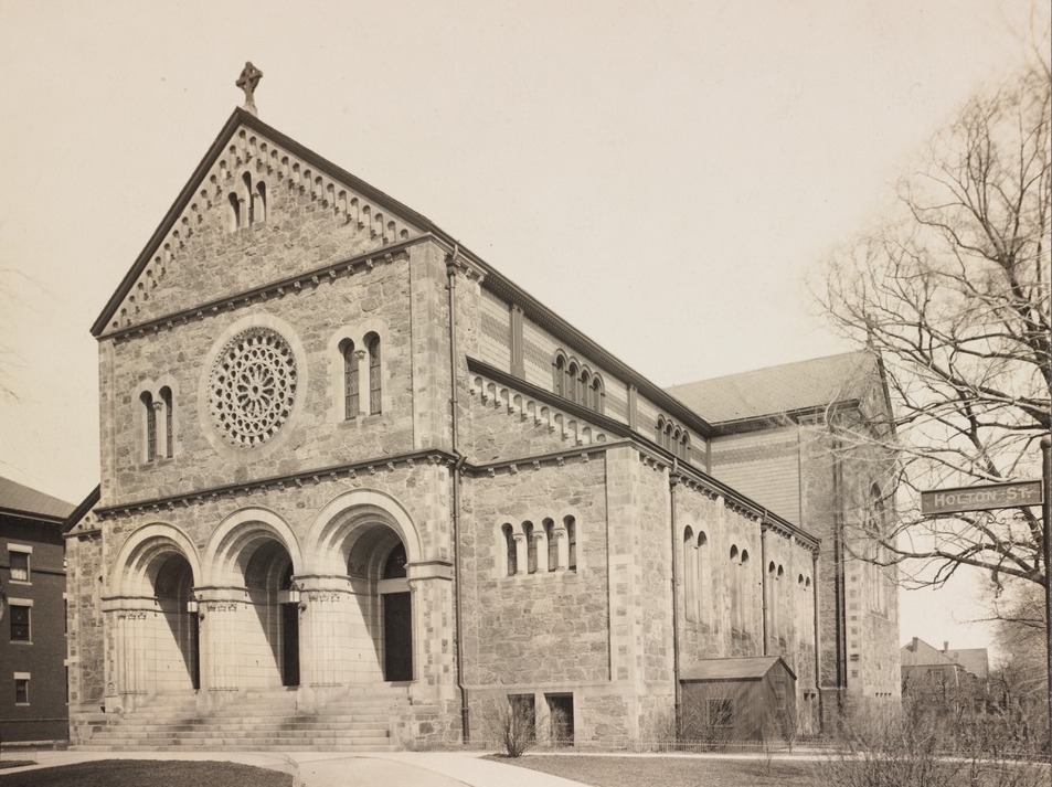A large church with three large arches at the entranceway. Throughout the building, there are many arched windows along with a large gothic rose window right above the entryways. The church is on Holton Street, as noted by a street sign on the left side.