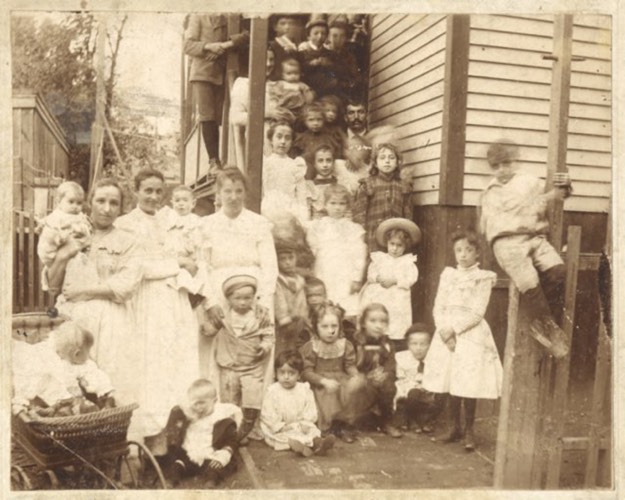 A photograph of a family seated on the steps and in front of their family home. There are many women, children, and babies in the photograph, with at least thirty-two people in the photograph.