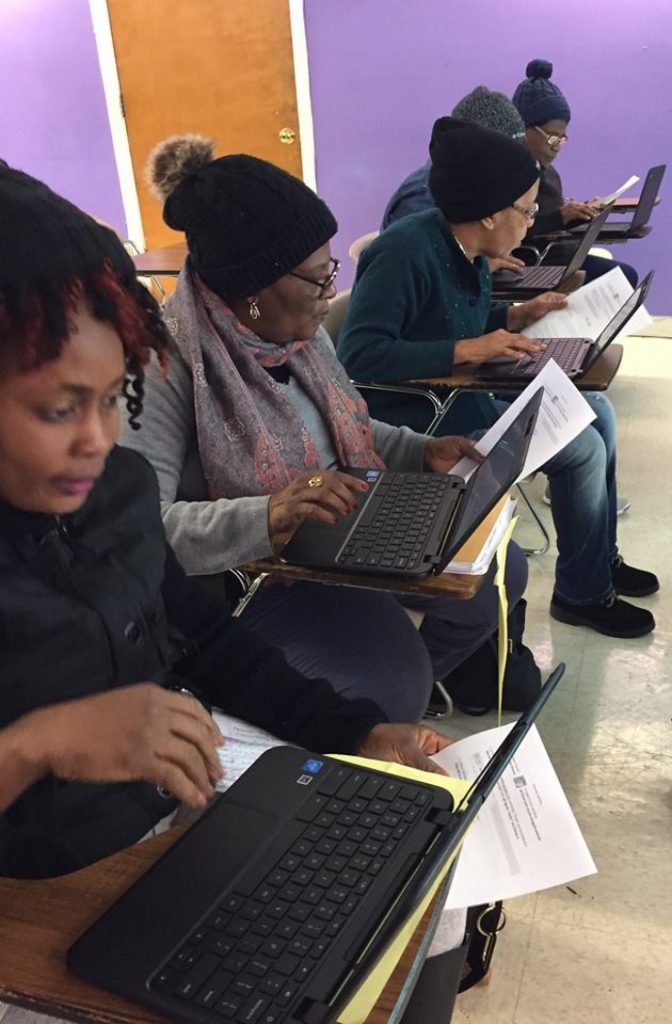 Four women sit at desks holding a piece of paper in one hand and have a laptop in front of them as they type and use the cursor with their other hand.