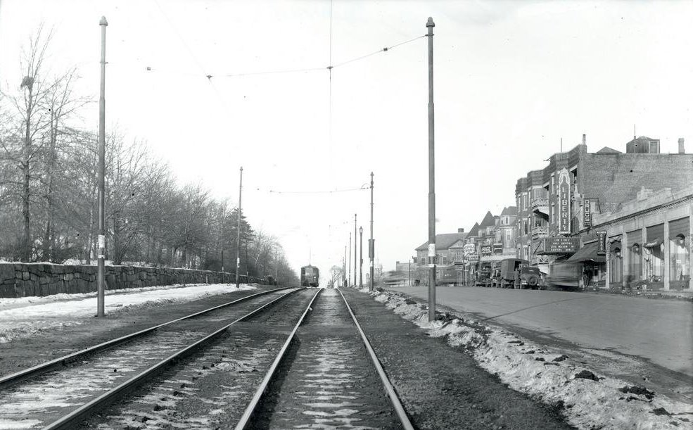 A photograph depicting streetcar lines on Blue Hill Avenue. A streetcar comes in the distance. On the righthand side of the photograph is a road with various buildings, businesses, and a few parked cars. On the left side of the photograph is a wooded area.