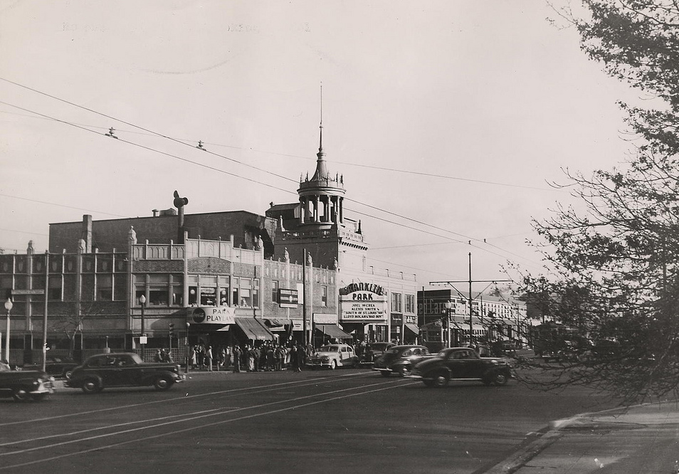 A photograph of the Franklin Park Theater, a large building that spans much of the street. On the street, cars pass by. There are many individuals in front of the theater, walking and waiting. In the middle of the theater, there is a tall spire.