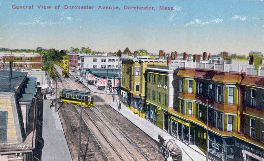 A postcard of Dorchester Avenue. A yellow streetcar travels down the street along with a horse-drawn wagon. On either side of the street are mixed-use buildings.