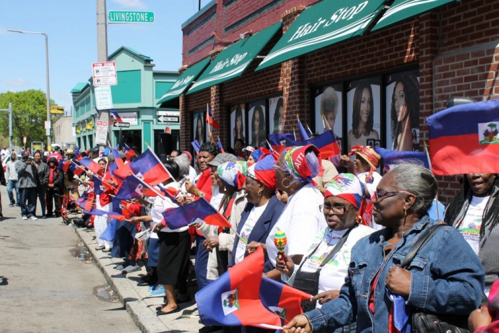 A sidewalk filled with many Haitian men and women waving hand-held Haitian flags. Many women wear depictions of the flag on their hair-coverings and many wear the same shirt for the parade.
