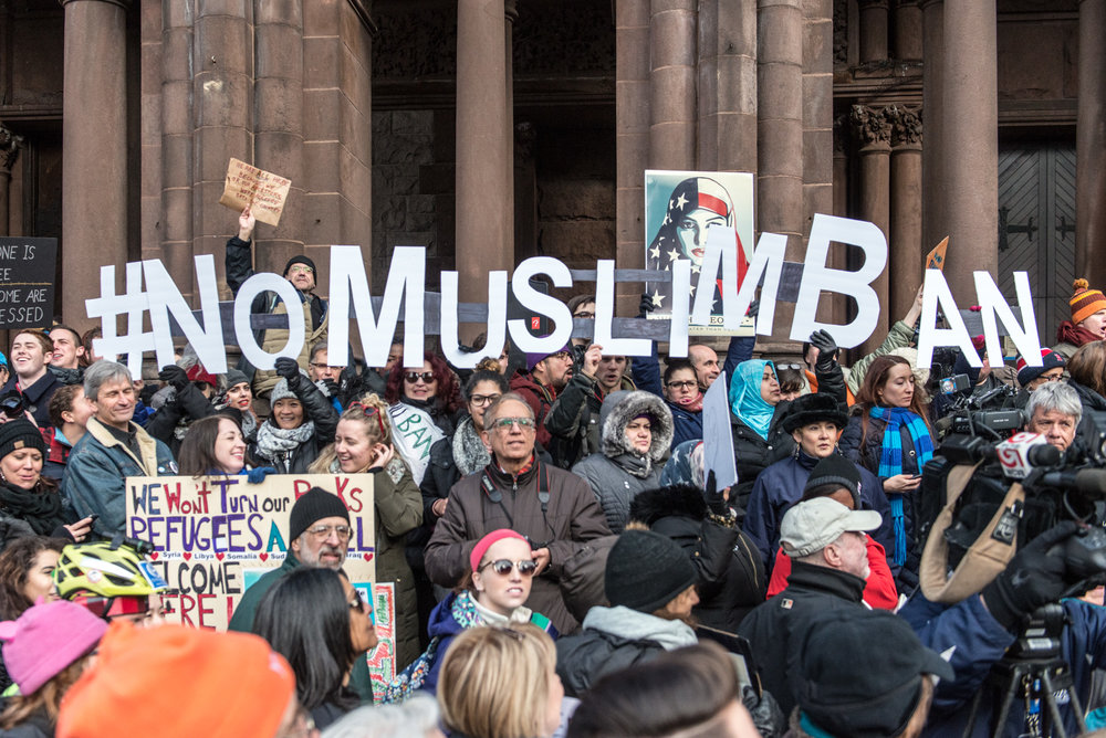 A photograph of many individuals protesting in their winter clothing. Many individuals hold various letters to spell out #No Muslim Ban. There are other various signs and posters throughout the photograph, such as one that says, "We won't turn our backs. Refugees are all welcome."