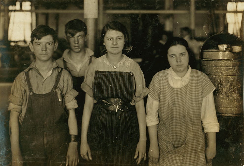 A photograph of two young boys in button-downs and overalls and two young girls in work smocks. The children are standing in front of the camera with the interior of the thread factory behind them.