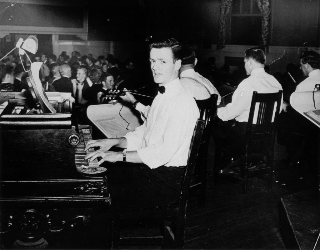 Frank Storer looks at the camera as he plays piano in the foreground. Behind him, there are three other instrumentalists, playing string instruments. In the background, there are many men and women dancing and talking.