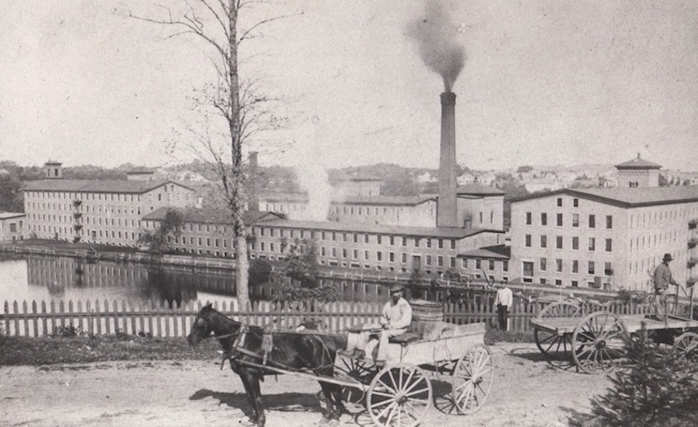 A photograph the mill behind a small body of water. The mill is quite long in length and at some points are six stories high. There are various buildings and two smoke-stacks. In the foreground of the photograph is a man driving a horse-drawn carriage.