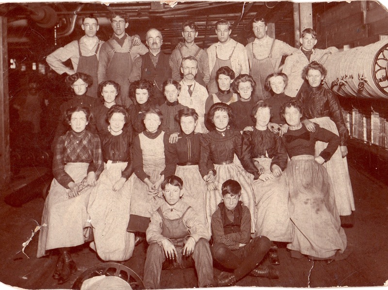 A photograph of workers at the mill. The workers are arranged into four rows. The first row shows two young boys seated, the second and third rows show fifteen women kneeling and standing. The back row shows nine men. The women all wear dark tops and lighter skirts. The men are all in overalls.