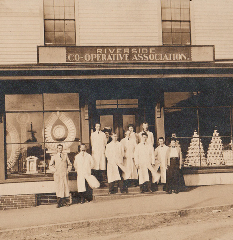 A photograph of eleven individuals in front of the Riverside Cooperative Association. The men all wear long white coats. The building has large storefront windows with displays on both sides.