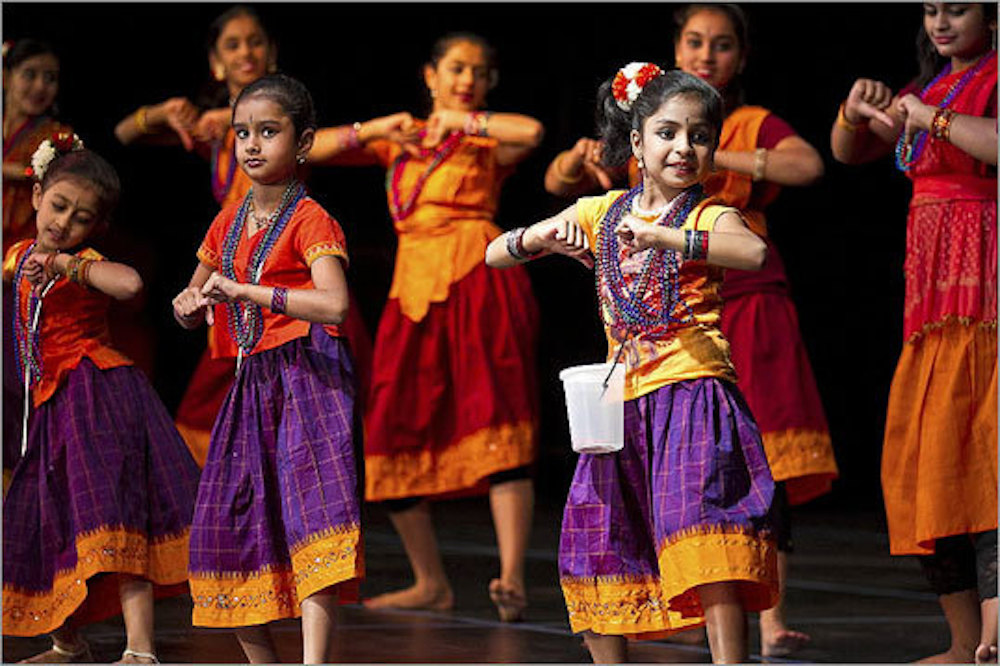 In the foreground, there are three young girls dancing in matching traditional Indian clothing, an orange short-sleeve shirt and a plaid purple and orange skirt. Behind the young girls is a row of older girls dancing as well.