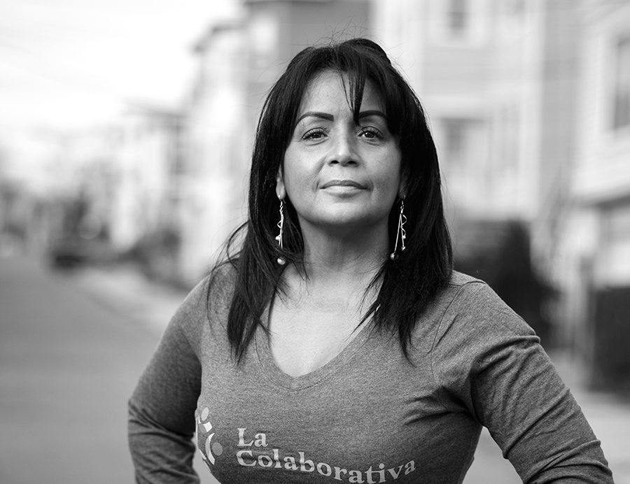 A photograph of a woman staring directly at the camera with her hands on her hip. She is wearing earrings and a long-sleeved shirt that says "La Colaborativa" on the chest. Behind the woman is a neighborhood street, although this is out of focus.