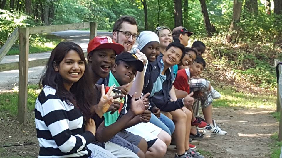 A photograph of eleven adolescents and youths holding hands on the side of the road in a wooded area. They are all smiling towards the camera.