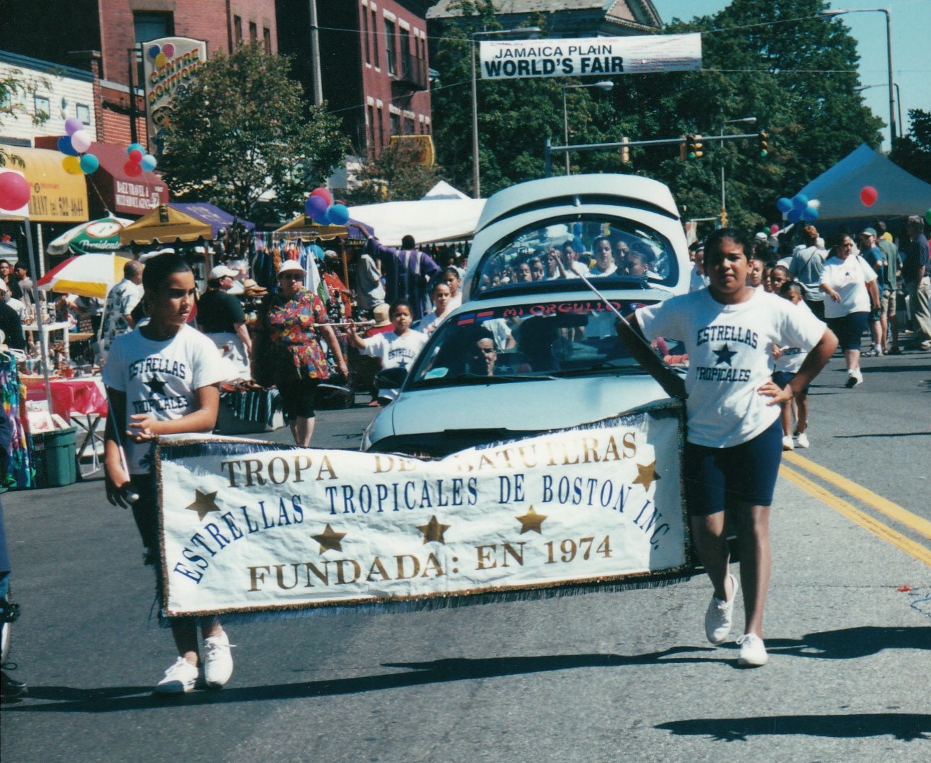 Two Youth Carrying A Banner In A Parade For The Jamaica Plain World's Fair, With A Bustling Street Fair In The Background.