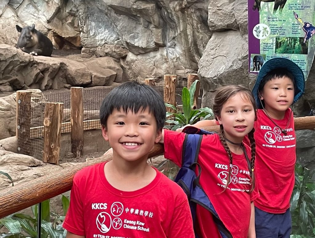 Three smiling young students wearing Kwong Kow T-shirts shown visiting a zoo.